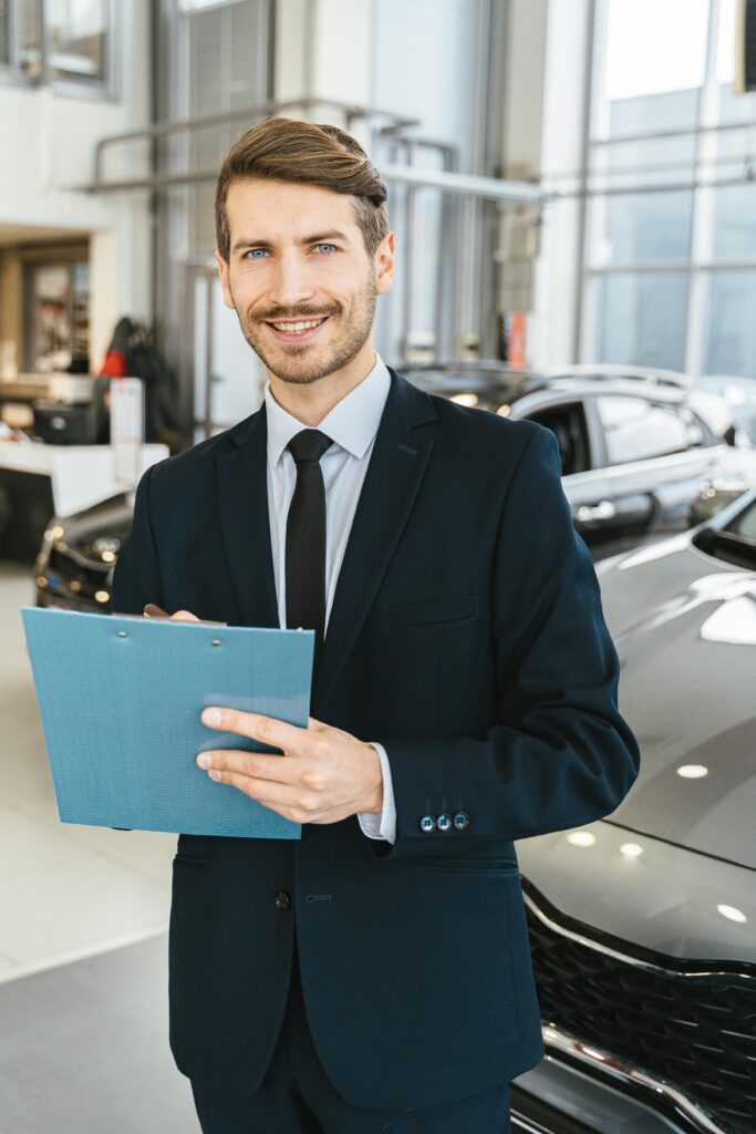 Confident car salesman in a suit with clipboard in a modern dealership showroom.